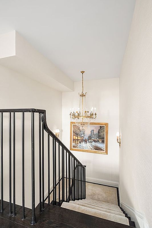 This image showcases an elegant staircase with dark wood steps and a black iron railing. A chandelier hangs above, illuminating a framed painting on the wall. The walls are painted in a neutral tone, creating a sophisticated and inviting atmosphere.