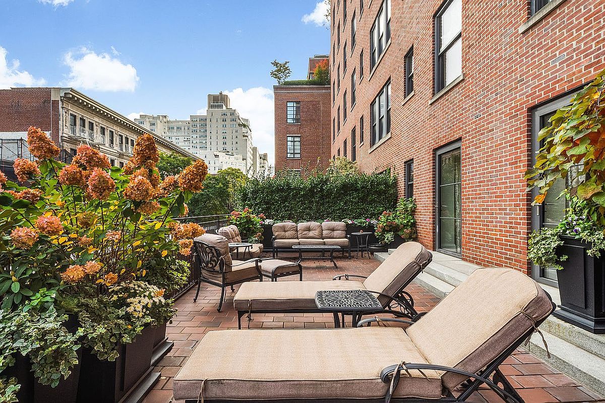 This image showcases a luxurious outdoor patio or balcony area, furnished with comfortable lounge chairs, a sofa, and tables, creating an inviting space for relaxation and entertainment. The patio is paved with brick and features lush greenery, including potted plants and a hedge, adding to the serene atmosphere. The backdrop includes a brick building and cityscape, suggesting an urban setting with a touch of nature.