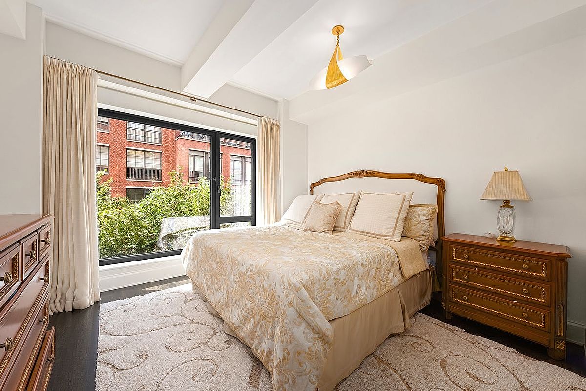 This is a cozy primary bedroom featuring a large window with a view of greenery and a red brick building. The room is furnished with a bed dressed in a patterned beige comforter and pillows, a wooden nightstand with a lamp, and a dresser. The overall style is traditional and inviting, with a focus on natural light and comfort.