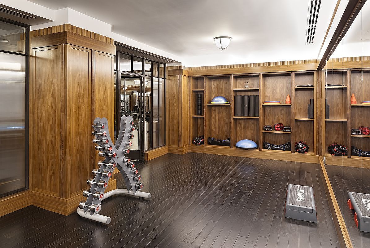 This is a well-appointed home gym featuring dark wood flooring and wood paneled walls with built-in shelving. A dumbbell rack with various weights is prominently displayed, along with a Reebok step platform. The room is well-lit and appears spacious, creating an inviting workout environment.