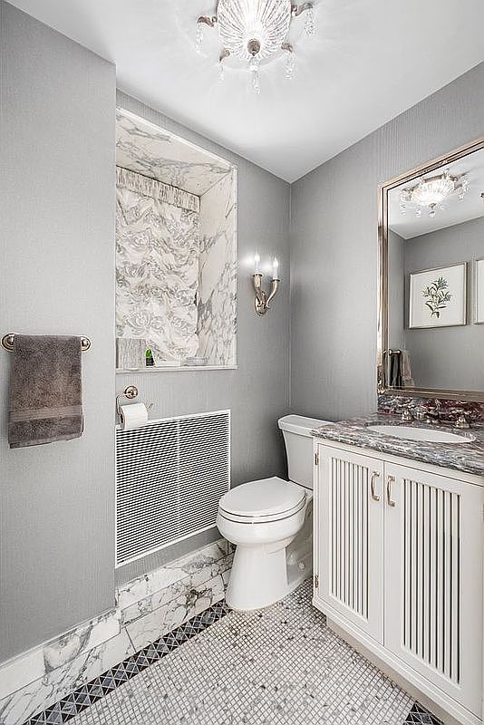 This is a well-appointed guest bathroom featuring a white toilet, a vanity with a marble countertop, and a framed mirror. The walls are painted in a neutral gray, and the floor is tiled with a mosaic pattern. A window with a marble frame and patterned curtain adds natural light and a touch of elegance.