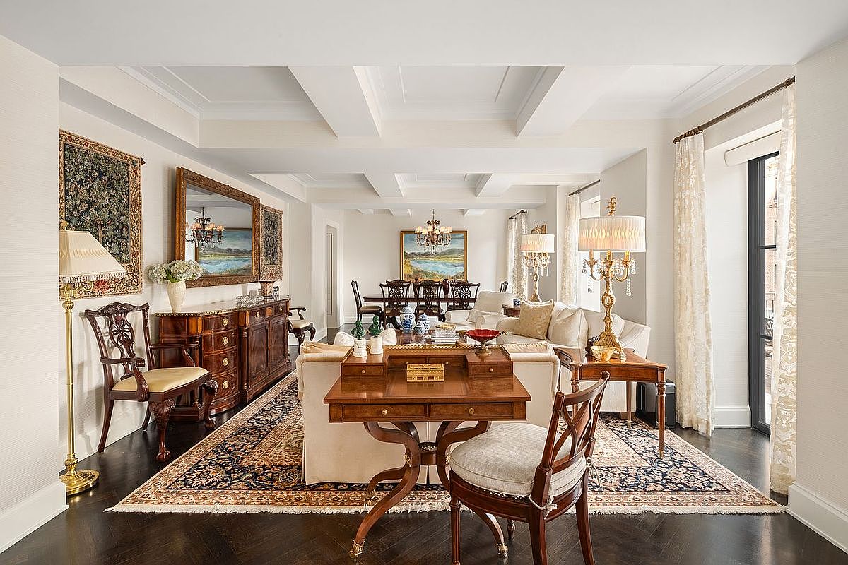 This is a traditionally styled living room featuring dark hardwood floors covered by an ornate area rug. The room includes a secretary desk, a sofa, and several antique-style chairs and lamps. The coffered ceiling and large windows add to the room's elegance and spaciousness.
