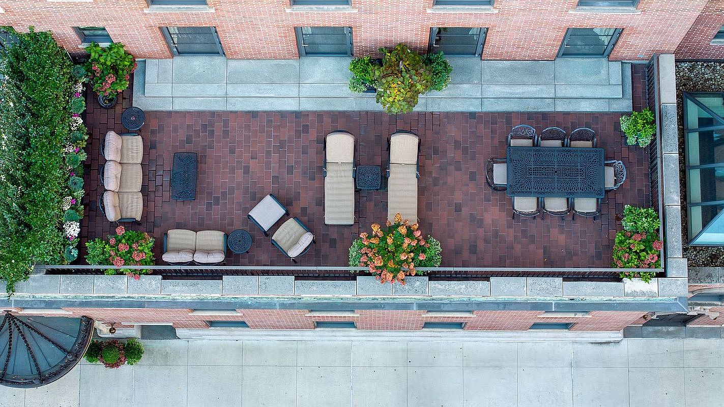 This is an aerial view of a luxurious rooftop patio. The patio features brick flooring, multiple seating areas with comfortable outdoor furniture, including sofas, lounge chairs, and a dining table set. Lush greenery and potted plants add a touch of nature to the urban setting, creating an inviting and relaxing outdoor space.