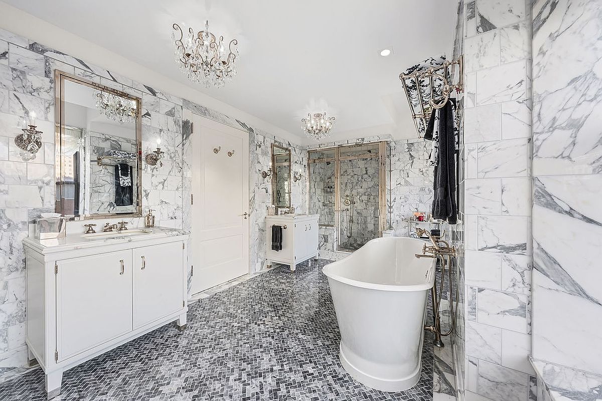 This is a luxurious primary bathroom featuring marble walls and a mosaic tile floor. The room includes a freestanding bathtub with gold fixtures, a glass-enclosed shower, and a vanity with a large mirror. Two elegant chandeliers provide ambient lighting, enhancing the bathroom's sophisticated and upscale ambiance.
