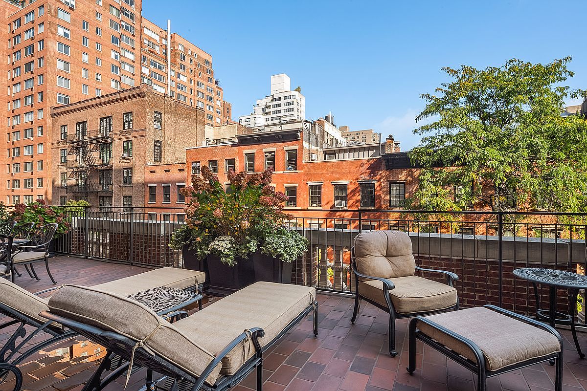 This image showcases a spacious outdoor patio or balcony area, furnished with lounge chairs, a small table, and a comfortable armchair with an ottoman. The patio features a brick floor and a black metal railing, offering a view of the surrounding cityscape with brick buildings and greenery. The scene suggests a relaxing and private outdoor living space.