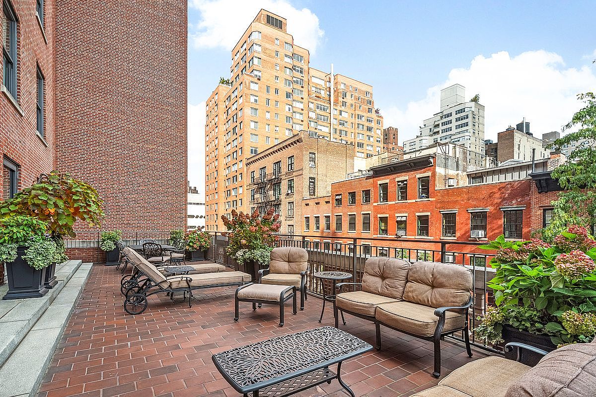 This image showcases a spacious outdoor patio or balcony area, furnished with comfortable seating arrangements including sofas, chairs, and chaise lounges. The patio is paved with red brick and features potted plants and flowers, adding a touch of greenery and color. The view extends to surrounding buildings, creating an urban oasis.