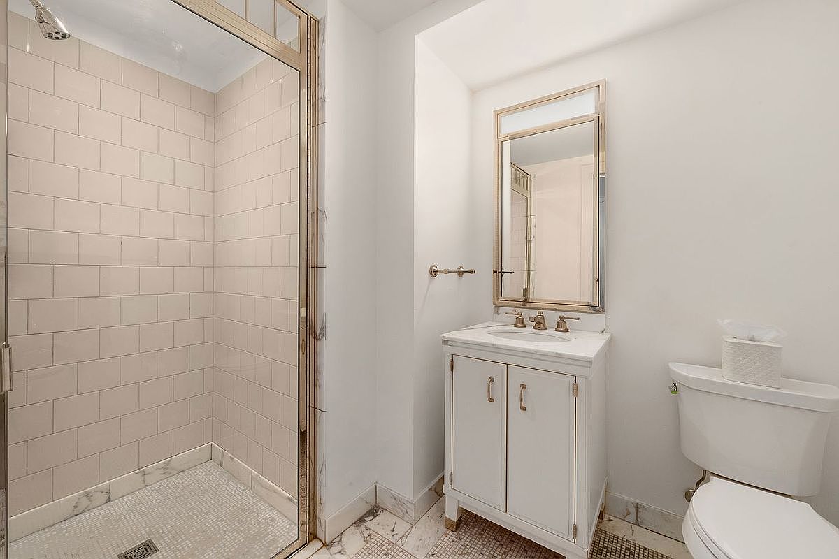This is a well-lit bathroom featuring a shower with glass doors and light pink tile, a white vanity with a marble countertop, and a matching framed mirror. A toilet is positioned next to the vanity. The bathroom has a clean and classic design, with marble flooring adding a touch of elegance.
