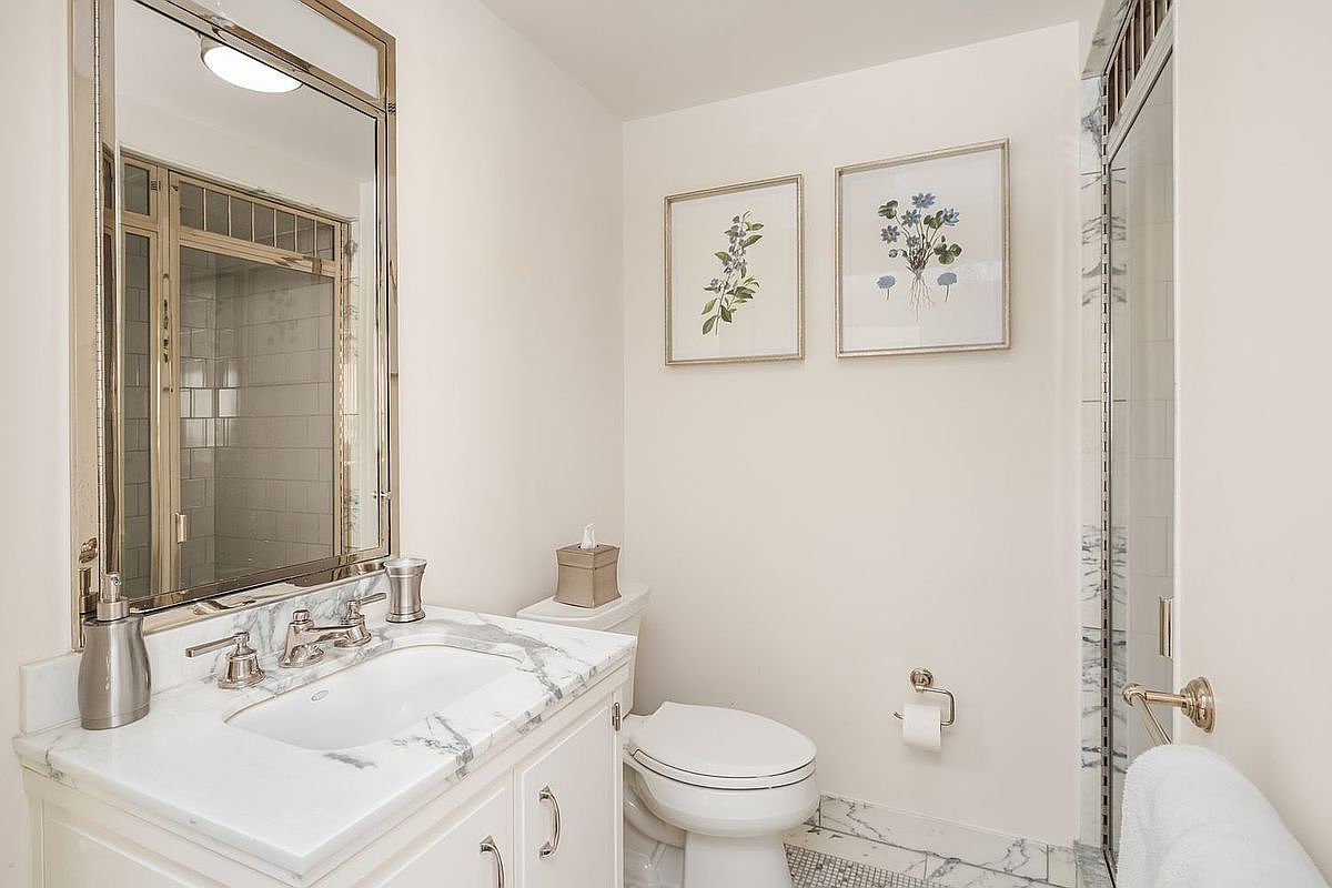 This is a well-lit bathroom featuring a white vanity with a marble countertop and a rectangular sink. A large mirror with a metallic frame hangs above the vanity, reflecting the shower area. Two framed botanical prints adorn the wall above the toilet, adding a touch of elegance to the space.