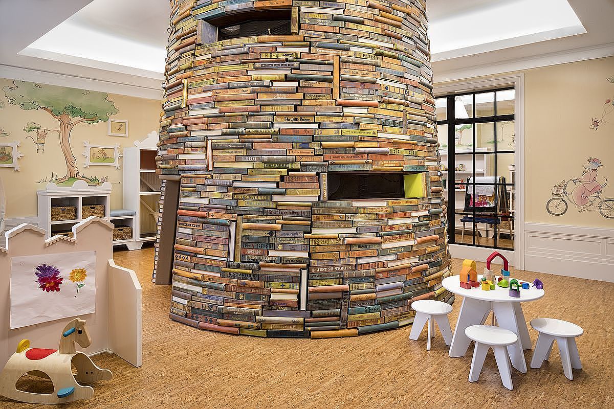 This is a whimsical children's playroom featuring a unique book tower as a central architectural element. The room is decorated with playful murals and includes a small table with stools, creating an inviting space for imaginative play. The overall impression is cheerful and creative, perfect for a young child's bedroom or playroom.