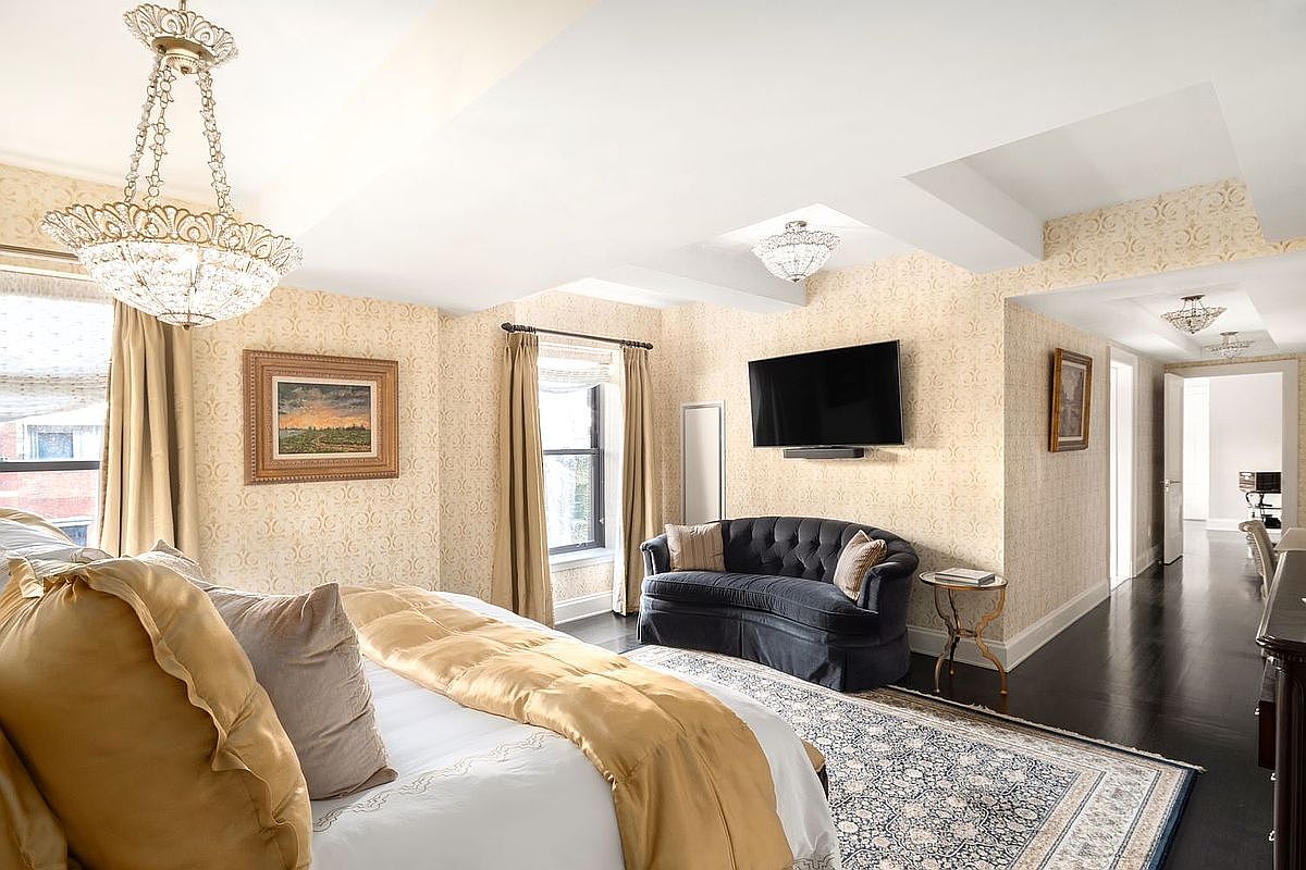 This is a luxurious primary bedroom featuring a large bed with gold accents, a tufted loveseat, and a wall-mounted television. The room is decorated with patterned wallpaper and elegant chandeliers, creating a sophisticated and comfortable atmosphere. A hallway leads to other parts of the home, showcasing the spacious layout.