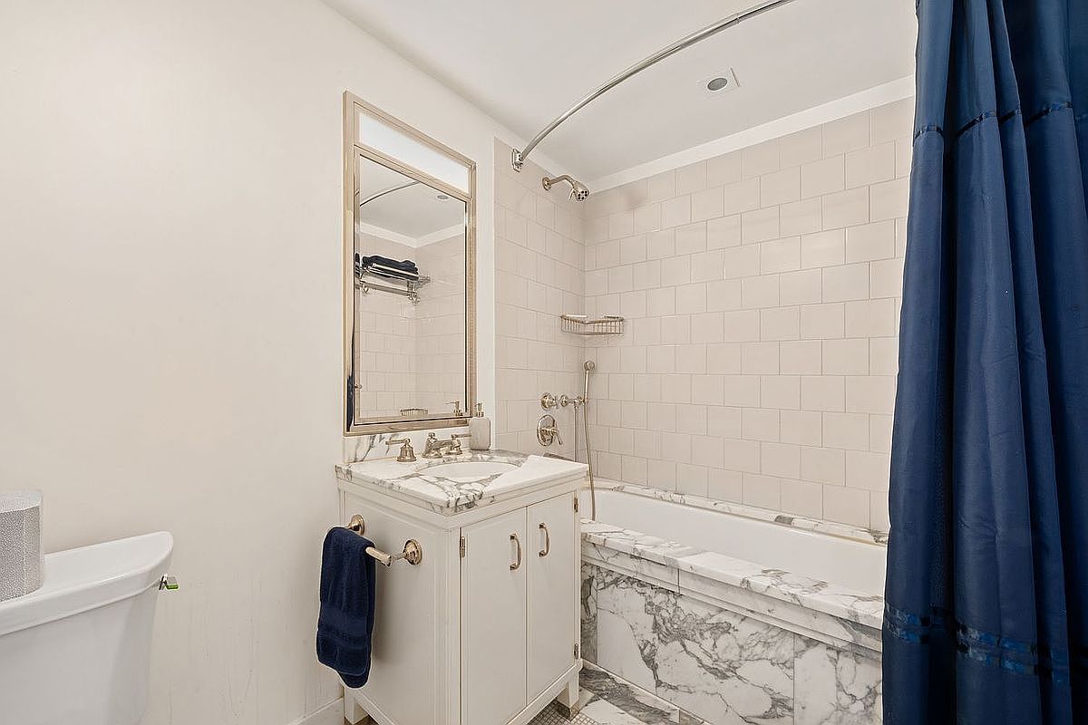 This is a bathroom featuring a white vanity with a marble countertop, a framed mirror, and a bathtub with white subway tile and a marble surround. A blue shower curtain is partially drawn, and a blue towel hangs on the vanity. The bathroom has a classic and clean aesthetic.
