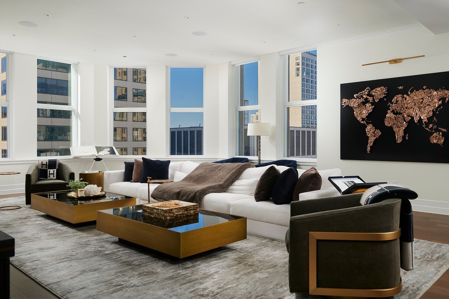 This sophisticated living room features a large, plush white sectional sofa accented with dark throw pillows and a cozy brown blanket, centered around two matching gold-framed coffee tables with dark glass tops. A striking, textured metallic world map art piece serves as a focal point on the wall, while floor-to-ceiling windows flood the space with natural light and offer urban city views. The room is anchored by a large, neutral-toned area rug and complemented by dark, modern armchairs, creating an atmosphere of refined luxury and comfort.
