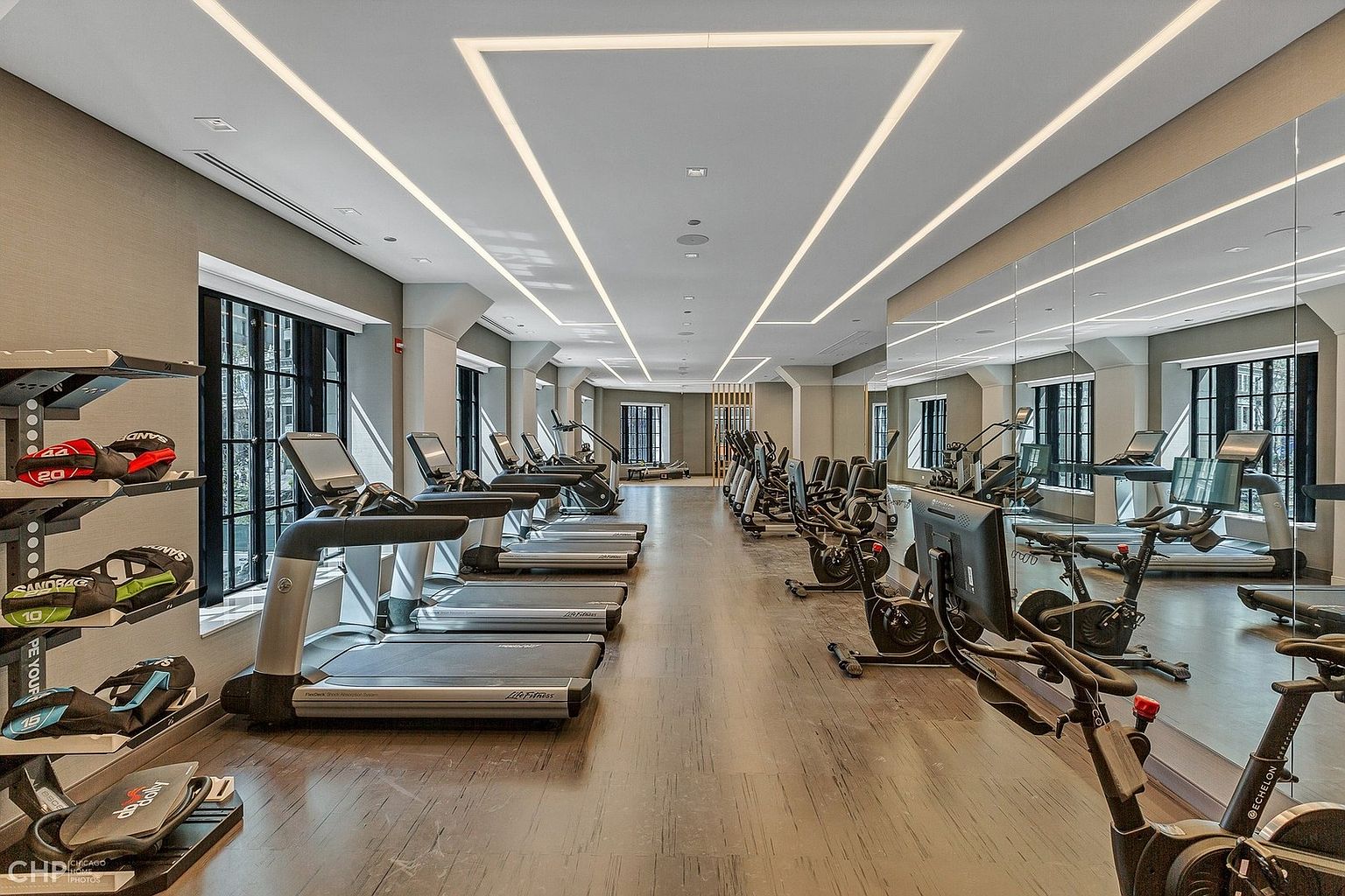 This spacious, modern fitness center features a long row of professional-grade treadmills and stationary bikes arranged neatly on a sleek wood-look floor. The room is illuminated by striking, geometric recessed ceiling lights and features a large wall-to-wall mirror that enhances the sense of depth and openness. A rack of weighted sandbags is positioned in the foreground, emphasizing the facility's comprehensive equipment offerings for residents.