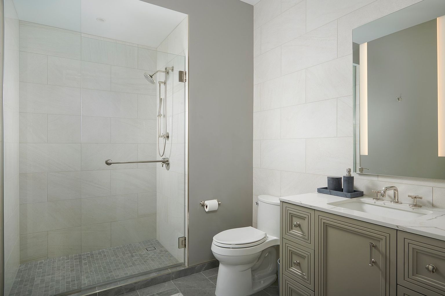 This modern, accessible bathroom features a walk-in shower with a glass enclosure, a grab bar, and light-colored tile walls. Adjacent to the shower is a white toilet and a vanity with a marble countertop, grey cabinetry, and a sleek mirror. The space is well-lit and clean, offering a functional and contemporary design.