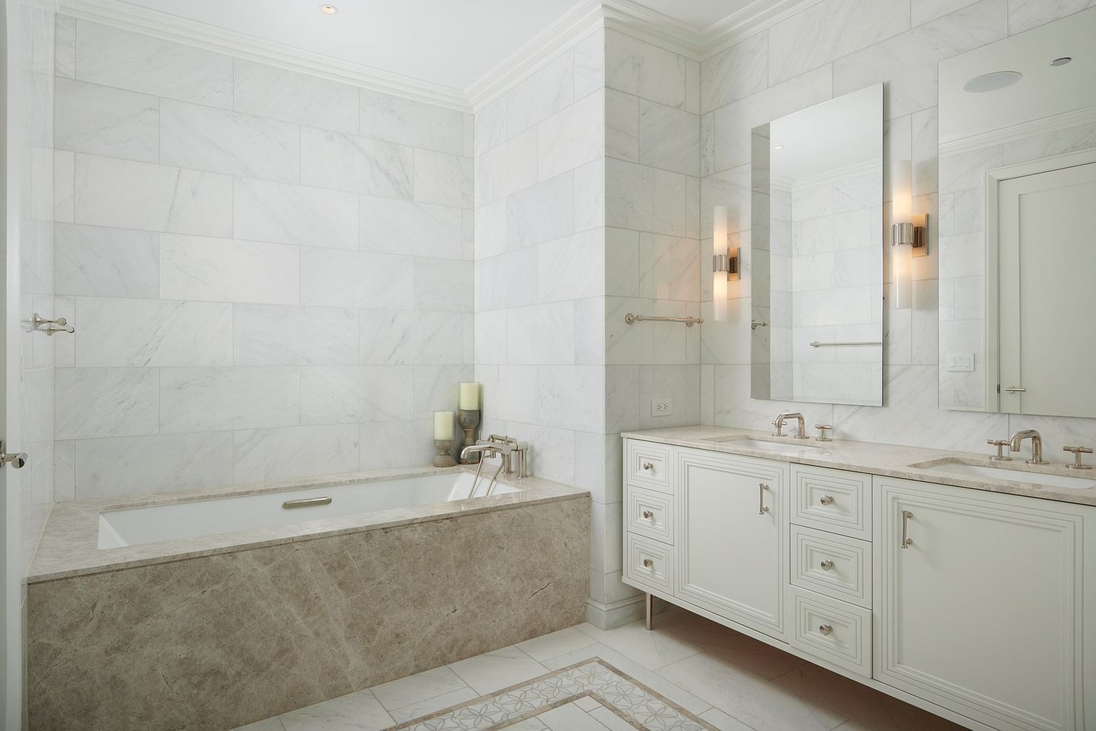 This elegant primary bathroom features a luxurious soaking tub encased in warm-toned stone, complemented by pristine white marble wall tiles. A double vanity with white cabinetry and matching stone countertops provides ample storage, while tall mirrors and sleek wall sconces add a sophisticated touch. The space is bright and airy, conveying a high-end, spa-like atmosphere perfect for a modern luxury home.