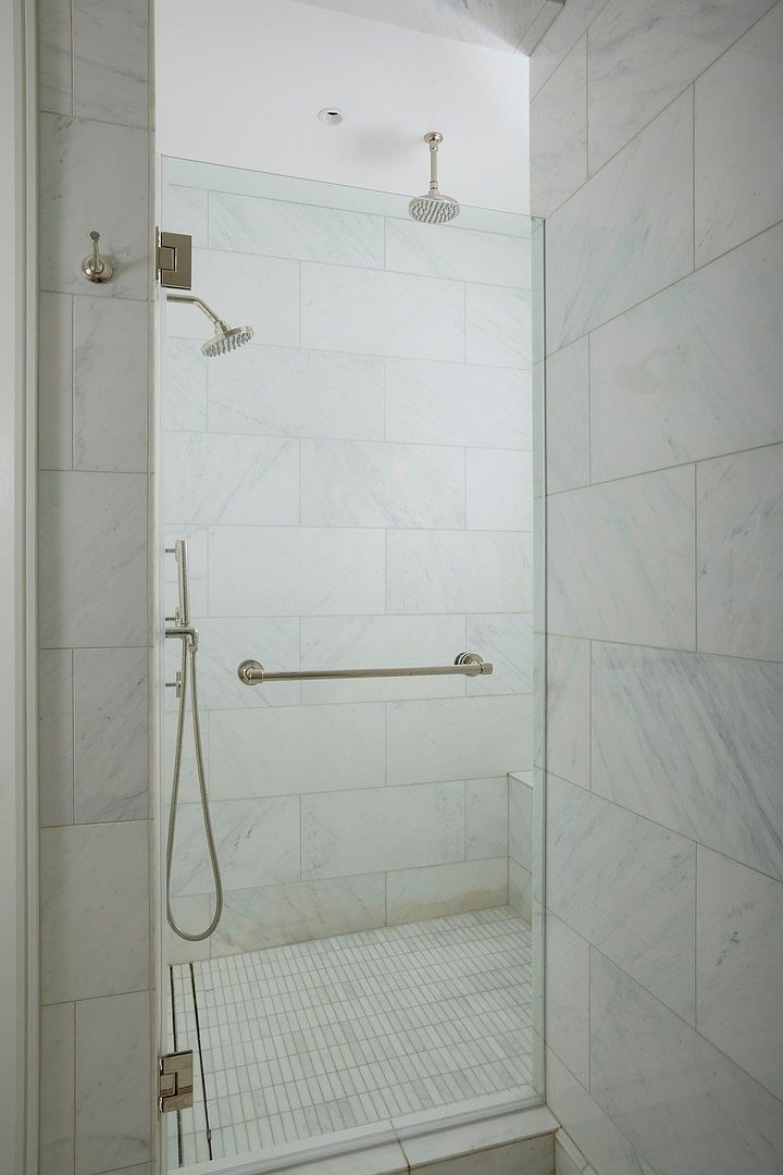 This modern walk-in shower features elegant white marble-look wall tiles and a clean, minimalist glass enclosure. It is equipped with both a ceiling-mounted rainfall showerhead and a separate handheld shower wand, along with a convenient grab bar for accessibility. The space is bright and airy, showcasing a high-end, functional design suitable for a luxury bathroom.