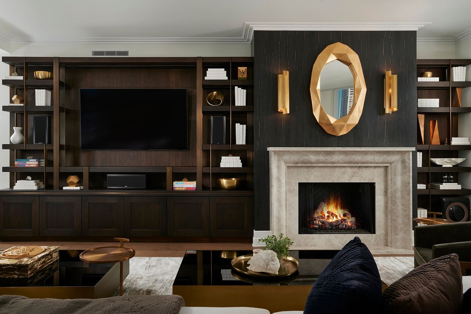 This sophisticated living room features a striking dark wood built-in media center and a grand stone fireplace as its focal points. The room is elegantly appointed with a geometric gold mirror, warm wall sconces, and a sleek black coffee table topped with decorative accents. The overall aesthetic is one of refined luxury, blending modern design with rich textures and warm, ambient lighting.