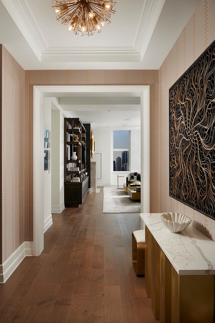 This elegant hallway features warm, textured wallpaper with vertical dotted patterns and rich hardwood flooring, leading the eye toward a bright, open living space. A sophisticated gold-toned console table with a marble top sits beneath a striking, intricate black and gold wall art piece, while a modern sputnik-style chandelier adds a touch of luxury to the ceiling. The perspective is centered and inviting, showcasing a seamless transition between the refined entryway and the sunlit living area beyond.