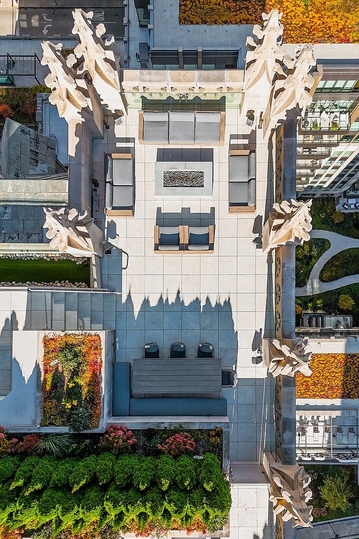 This stunning aerial view captures a luxurious rooftop terrace featuring a sophisticated outdoor living space framed by ornate Gothic-style architectural stone carvings. The layout includes a cozy lounge area with a central fire pit, a dining section with a long table, and lush, manicured garden beds that add vibrant color to the stone patio. The perspective provides a cinematic look at an exclusive urban oasis, highlighting both the high-end furnishings and the intricate historic-inspired design elements.