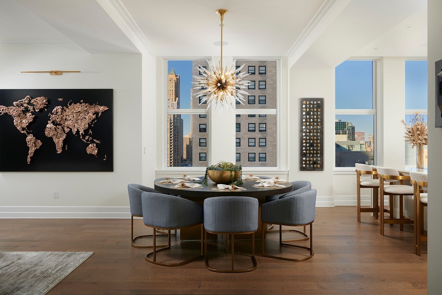 This sophisticated dining room features a large, dark circular table surrounded by six plush, blue-upholstered chairs with gold metal bases. A striking starburst chandelier hangs above the table, serving as a focal point, while a large, textured metallic world map art piece adorns the wall. The room is bathed in natural light from expansive windows that offer a clear view of the surrounding city skyline, creating an elegant and modern urban atmosphere.