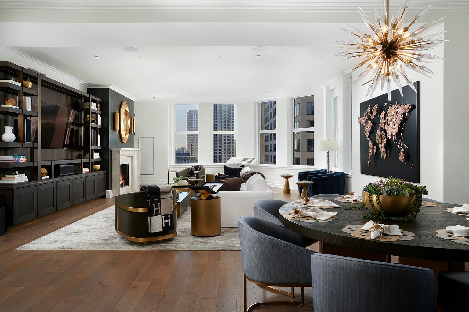 This expansive, open-concept living and dining area exudes modern luxury with its sophisticated dark wood cabinetry, plush white sofa, and high-end designer accents. A striking starburst chandelier hangs above a circular dining table, while a large, textured world map art piece serves as a focal point on the wall. The space is bathed in natural light from floor-to-ceiling windows, offering a seamless blend of comfort and high-style urban living.