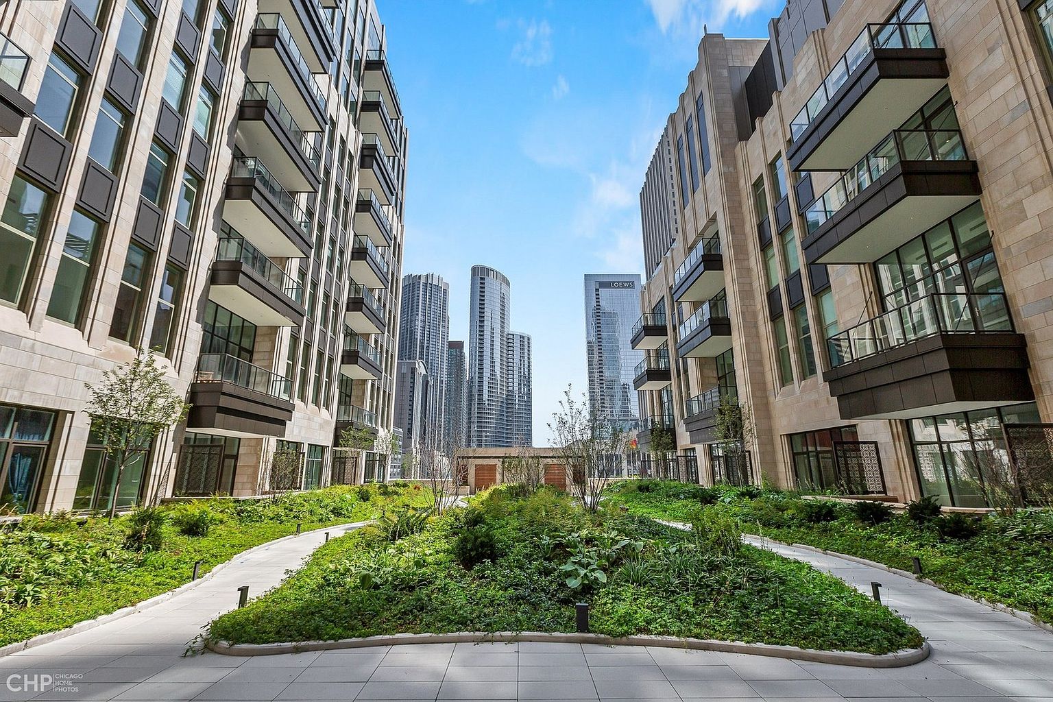 This image captures a beautifully landscaped courtyard garden nestled between two modern, high-rise residential buildings. The space features winding stone pathways, lush greenery, and small trees, creating a serene urban oasis. In the background, the iconic Chicago skyline, including the Loews hotel, rises above the courtyard, providing a striking contrast between nature and city architecture.