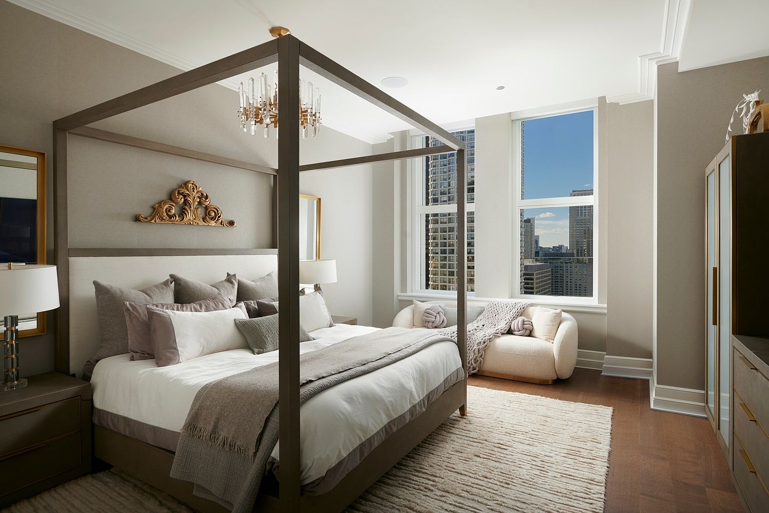 This elegant primary bedroom features a sophisticated four-poster bed as the centerpiece, adorned with plush neutral-toned bedding and a decorative gold wall accent. A cozy, curved cream-colored loveseat sits by the large windows, which offer a stunning urban skyline view, while a textured area rug adds warmth to the hardwood floors. The room exudes a luxurious, serene atmosphere with its refined color palette and high-end finishes.
