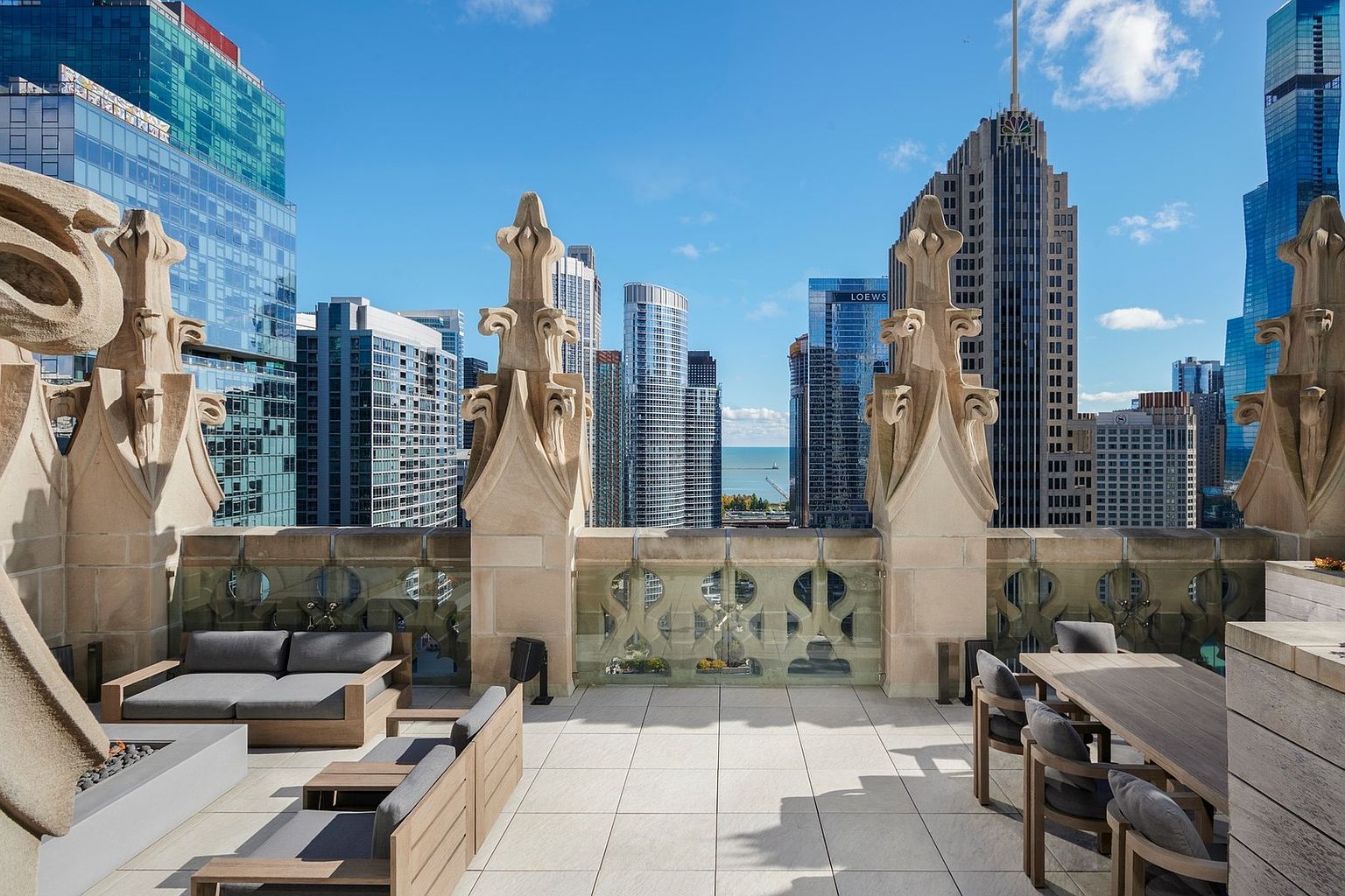 This luxurious rooftop terrace offers a sophisticated outdoor living space with stunning panoramic views of the Chicago skyline and Lake Michigan. The area is furnished with modern, comfortable seating and a dining set, framed by ornate, gothic-inspired stone architectural details. The clean, paved flooring and glass railings create a seamless transition between the elegant terrace and the dramatic urban backdrop.