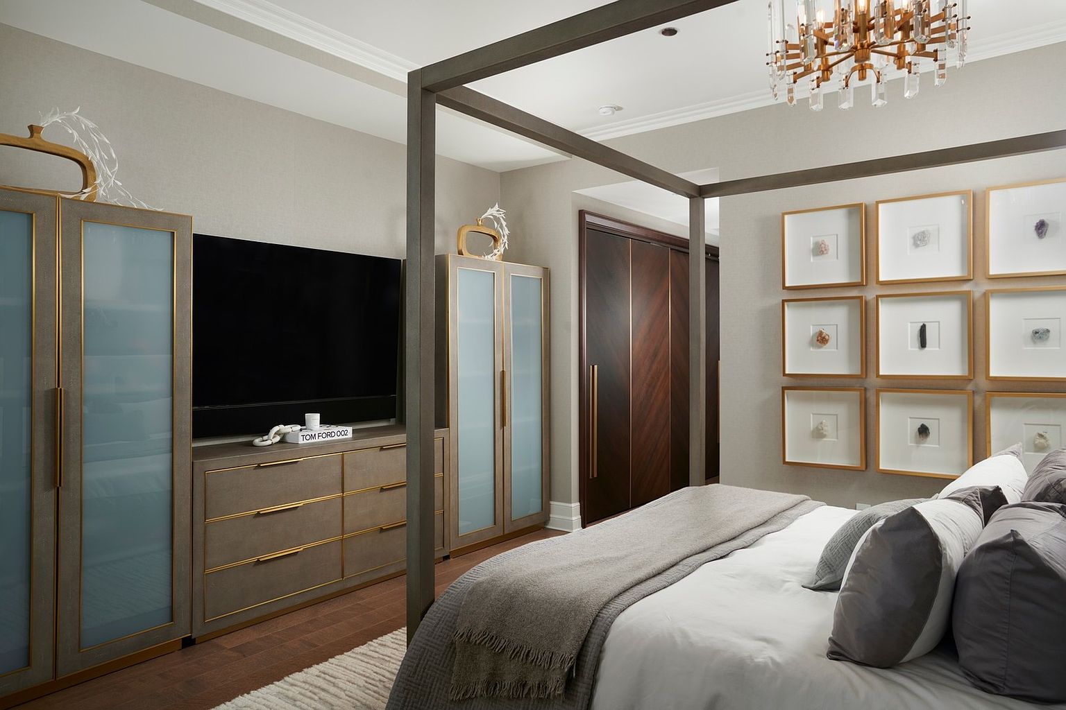 This sophisticated primary bedroom features a striking four-poster bed frame and a neutral, monochromatic color palette that exudes luxury. The room is accented by a sleek media console with a mounted television, flanked by two tall, glass-fronted cabinets with gold trim, and a gallery wall of framed mineral specimens. The warm wood flooring and soft, layered bedding create an inviting and high-end atmosphere suitable for a modern, upscale residence.