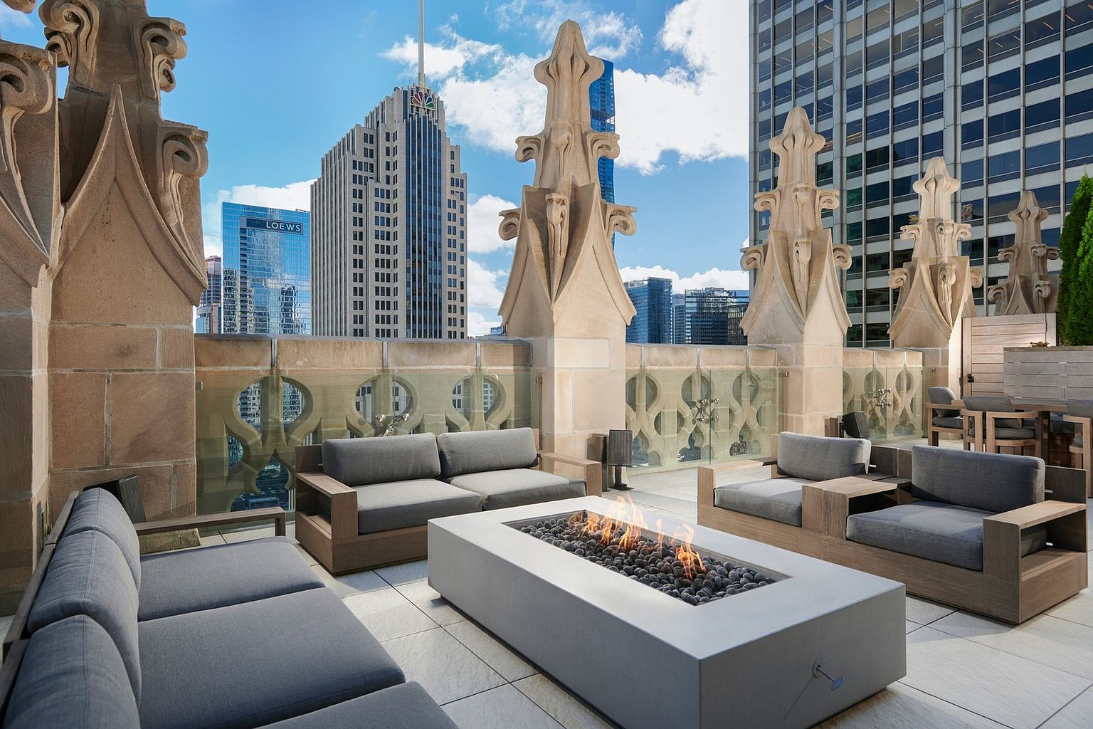 This luxurious rooftop terrace offers a sophisticated outdoor living space featuring modern, low-profile lounge seating arranged around a sleek, rectangular concrete fire pit. The area is framed by ornate, gothic-inspired stone architectural details and a glass railing that provides unobstructed, panoramic views of the surrounding city skyline. The combination of high-end contemporary furniture and historic-style masonry creates an elegant, upscale atmosphere perfect for urban entertaining.
