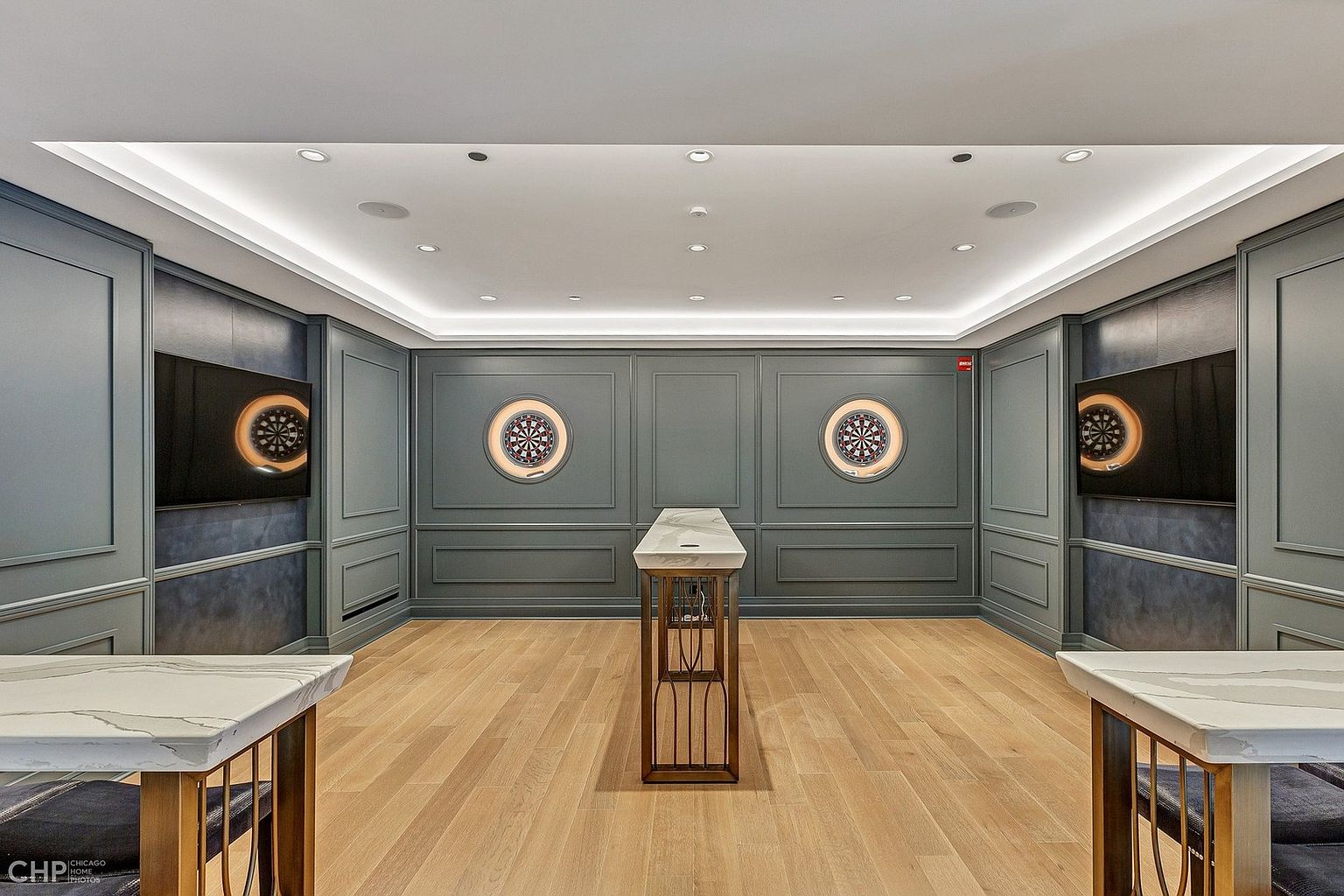 This sophisticated game room features a symmetrical layout with two dartboards mounted on a paneled, dark-toned wall, flanked by large flat-screen televisions. A central marble-topped console table anchors the space, complemented by matching side tables in the foreground, all set against light hardwood flooring. The room is illuminated by recessed lighting and a cove ceiling, creating a refined and modern atmosphere perfect for entertainment.