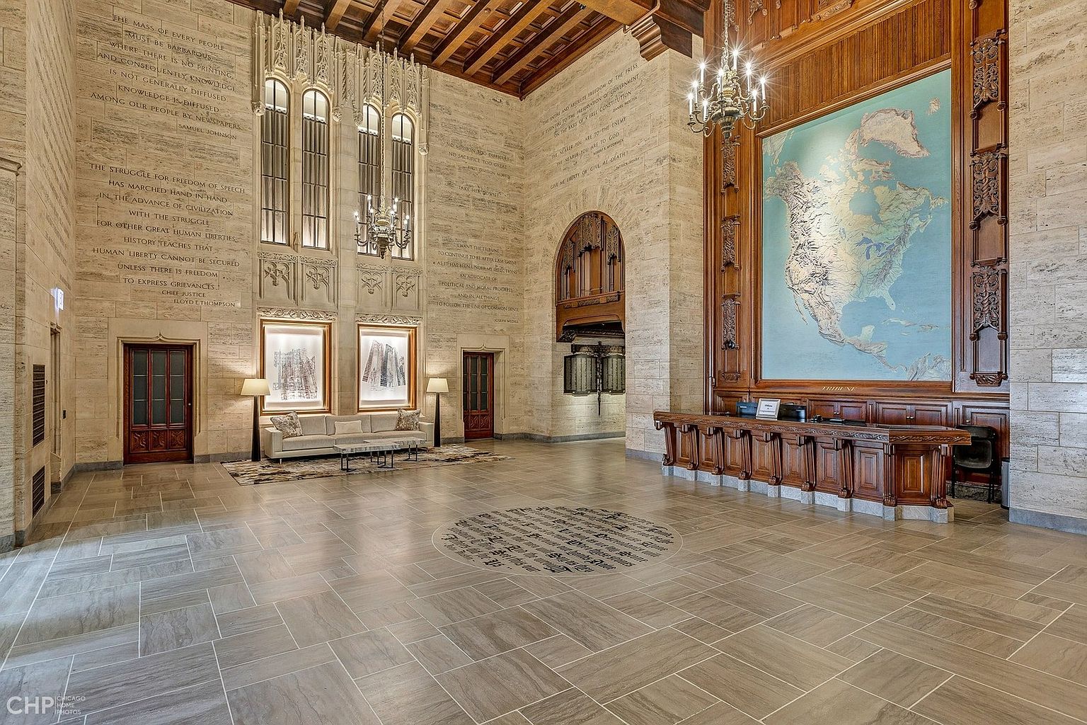 This grand, historic lobby features soaring ceilings, intricate stone carvings, and walls adorned with engraved literary quotes, evoking a sense of timeless prestige. A large, detailed map of North America serves as a focal point behind a long, dark wood reception desk, while a plush sofa provides a comfortable seating area. The perspective is a wide-angle shot that captures the immense scale and architectural elegance of this iconic building's entrance.