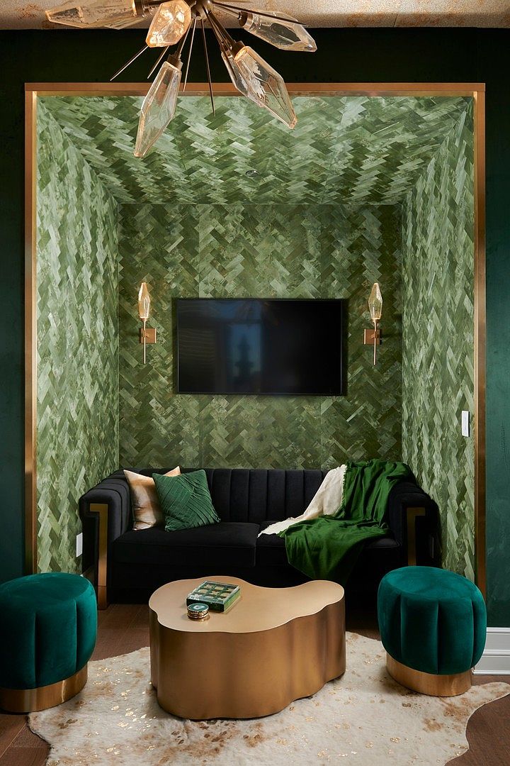 This sophisticated living space features a dramatic, recessed alcove lined with green herringbone-patterned wallpaper, framing a wall-mounted television and flanked by elegant gold sconces. A plush black velvet sofa sits atop a cowhide rug, complemented by two matching emerald green velvet ottomans and a sculptural gold coffee table. The room is illuminated by a striking, modern crystal chandelier, creating a luxurious and moody atmosphere perfect for a high-end lounge or media room.