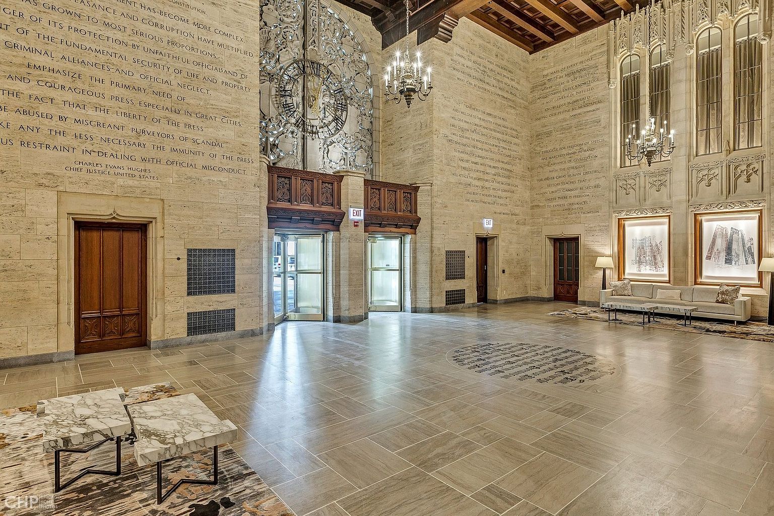 This grand, historic lobby features soaring ceilings, intricate stone carvings, and walls adorned with engraved historical quotes, creating an atmosphere of immense prestige and architectural significance. The space is anchored by a polished stone floor, elegant chandeliers, and a sophisticated seating area, offering a dramatic and welcoming entrance for residents or visitors. The perspective is a wide-angle shot from the center of the room, emphasizing the scale and opulent design of the building's common area.