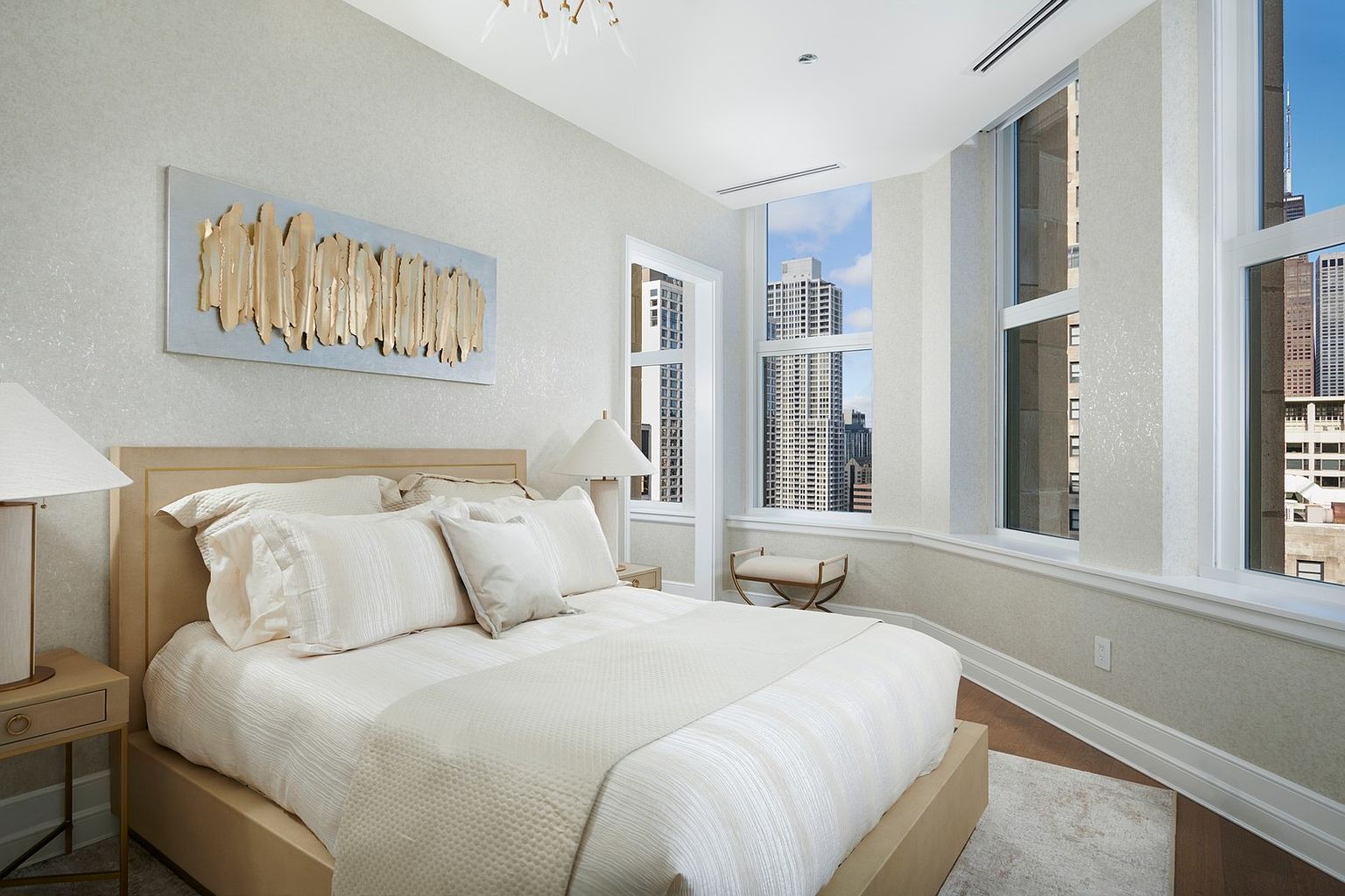 This elegant primary bedroom features a neutral color palette with a plush, upholstered bed as the focal point, adorned with textured linens and pillows. A striking gold-leaf abstract art piece hangs above the headboard, while large windows offer expansive urban views and flood the room with natural light. The space is finished with sophisticated bedside tables, a sleek bench, and a modern chandelier, creating a serene and luxurious atmosphere.