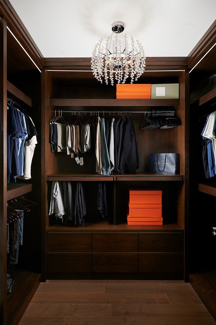 This sophisticated walk-in closet features rich, dark wood cabinetry and shelving, providing a luxurious and organized storage solution. A central crystal chandelier adds a touch of elegance, while the symmetrical layout and integrated lighting highlight the neatly arranged clothing and accessories. The overall aesthetic is refined and high-end, perfect for a modern, upscale home.