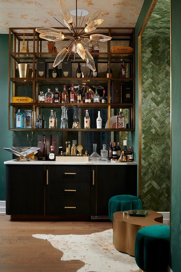 This sophisticated home bar area features a custom-built, floor-to-ceiling brass shelving unit filled with premium spirits and decorative accents, set against a dark, moody backdrop. Below, a sleek black cabinet with gold hardware provides ample storage, complemented by a plush, emerald green velvet stool and a unique, organic-shaped coffee table on a cowhide rug. The space is illuminated by a striking, modern starburst chandelier, creating an atmosphere of luxury and refined entertainment.