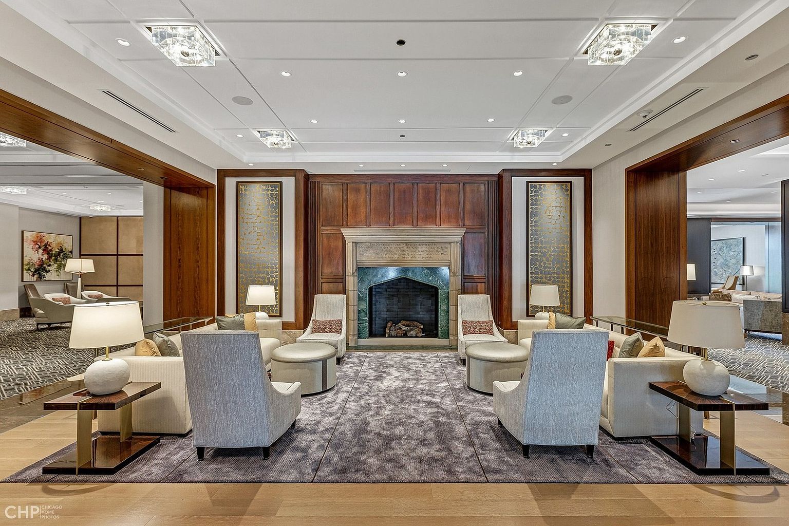 This elegant, symmetrical lounge area features a grand stone fireplace as its focal point, flanked by decorative gold-toned panels and rich wood-paneled walls. The space is furnished with plush, light-colored armchairs and sofas arranged on a large, textured area rug, creating a sophisticated and inviting atmosphere. The perspective is centered and eye-level, emphasizing the formal, high-end design of this communal space.