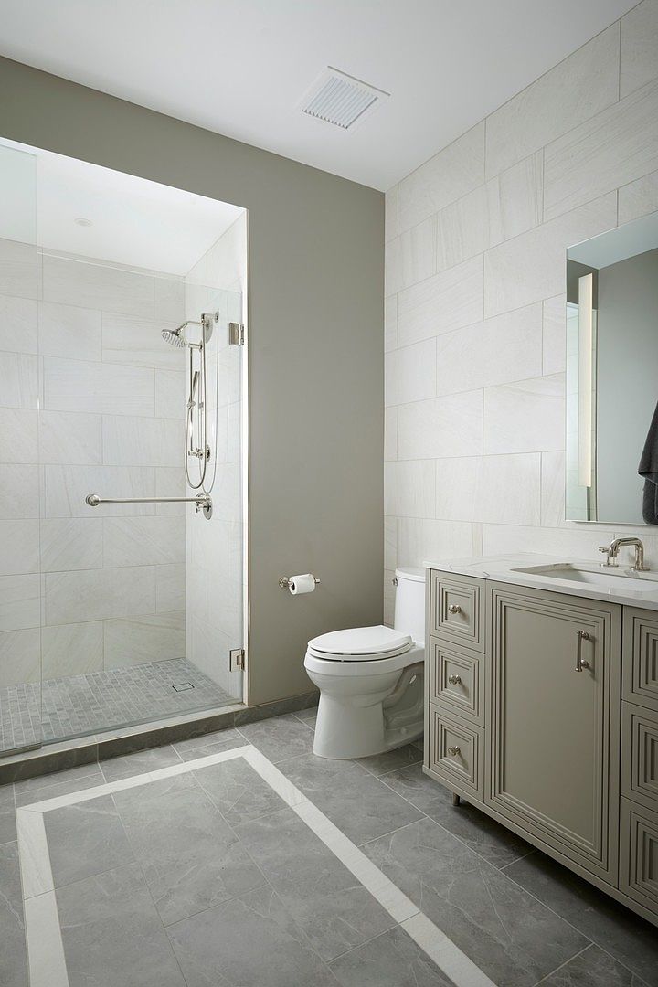 This modern primary bathroom features a sleek walk-in glass shower with light-toned tile and a separate vanity area with a neutral-colored cabinet and marble countertop. The floor is adorned with large-format grey tiles featuring a decorative white border, creating a sophisticated and clean aesthetic. The perspective is a wide-angle shot from the doorway, capturing the functional layout and contemporary design elements of the space.