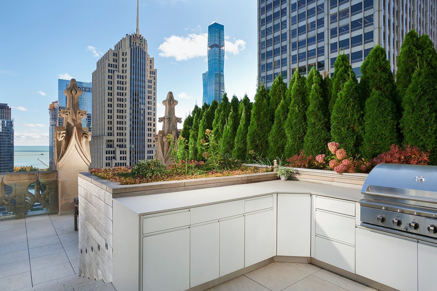 This high-end rooftop terrace features a sleek, modern outdoor kitchen with white cabinetry and a stainless steel grill, set against a backdrop of historic gothic-style stone architecture. The space is framed by a lush row of tall evergreen trees and offers a dramatic, panoramic view of the city skyline and the distant lake. The combination of contemporary amenities and classic architectural details creates a sophisticated and luxurious urban outdoor living environment.