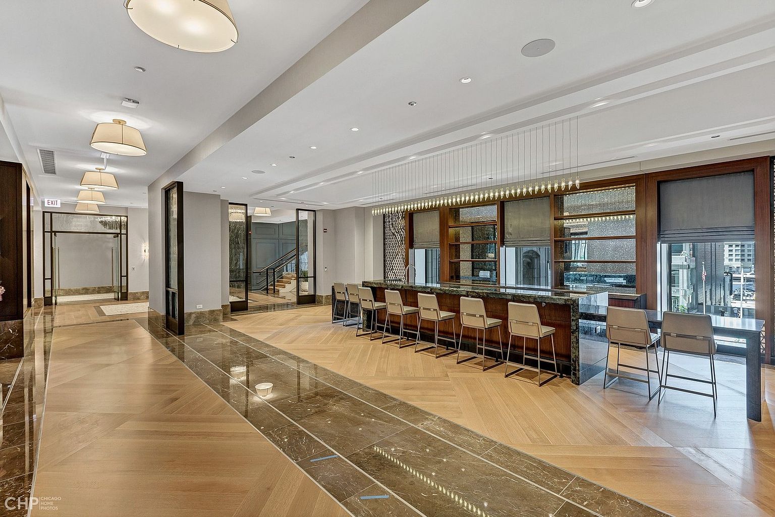 This sophisticated communal lounge area features a long, sleek bar with marble countertops and modern bar stools, set against a backdrop of warm wood paneling and large windows. The space is defined by a striking contrast between the herringbone-patterned light wood flooring and a central inlay of polished dark stone, creating a luxurious and inviting atmosphere. The perspective is a wide-angle shot from the hallway, emphasizing the expansive, high-end design and the elegant lighting fixtures that illuminate the room.