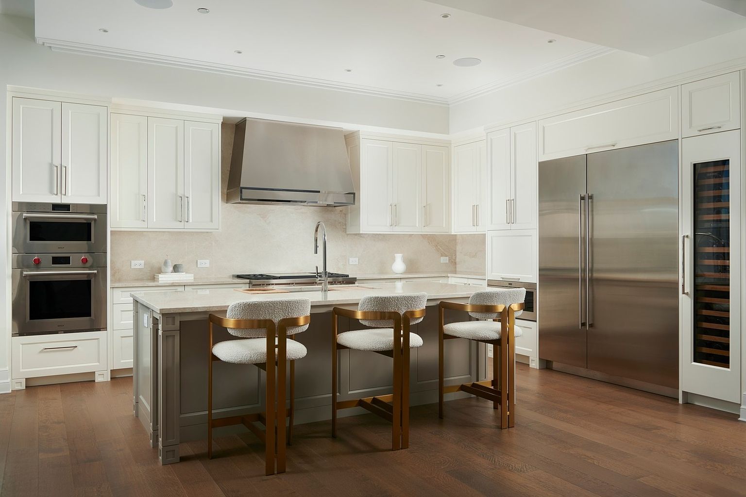 This sophisticated, high-end kitchen features a clean, transitional design with crisp white cabinetry, a large central island with a grey base, and warm-toned hardwood flooring. The space is equipped with professional-grade stainless steel appliances, including a double wall oven and a large refrigerator, complemented by elegant gold-accented bar stools. The perspective is a wide, eye-level shot that highlights the seamless integration of luxury finishes and functional layout, creating an inviting and polished atmosphere.