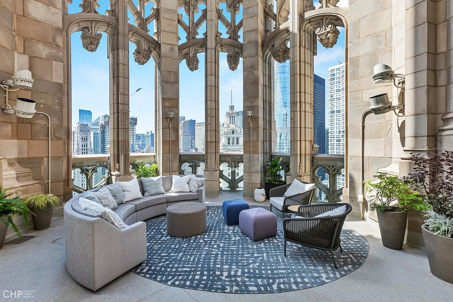 This stunning, high-rise balcony features intricate Gothic-style stone arches that frame panoramic views of the Chicago skyline. The space is furnished with a large, curved grey sofa, woven armchairs, and colorful ottomans arranged on a patterned circular rug, creating an elegant outdoor lounge area. The combination of historic architectural detailing and modern luxury furnishings offers a sophisticated and unique urban retreat.