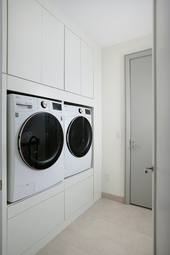 This clean and modern laundry room features a built-in, floor-to-ceiling white cabinetry system that seamlessly integrates a side-by-side LG washer and dryer. The space is finished with light-colored tile flooring and a neutral grey door, creating a bright, organized, and functional utility area. The perspective is a straight-on shot that highlights the efficient use of space and contemporary design.