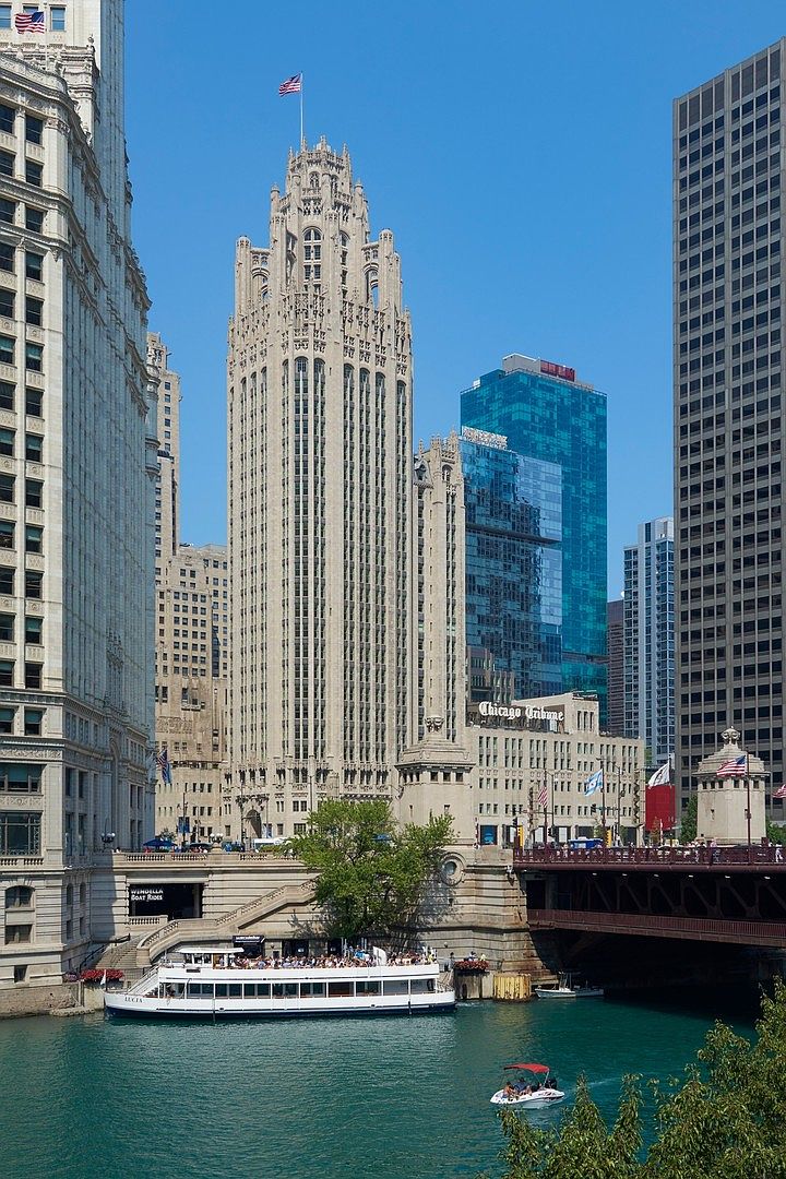 This iconic view captures the historic Tribune Tower in Chicago, showcasing its intricate neo-Gothic architecture and soaring spires against a clear blue sky. The scene includes the Chicago River in the foreground with a tour boat and a small motorboat, framed by the surrounding urban skyline and a bridge. The perspective is a wide, eye-level shot that emphasizes the grandeur and architectural significance of this landmark building within the city's bustling riverfront.