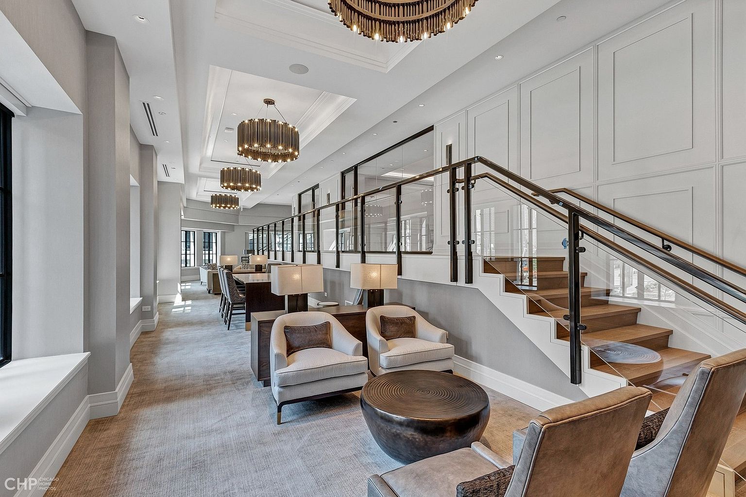This sophisticated, expansive lounge area features a modern aesthetic with high ceilings, elegant drum chandeliers, and a grand staircase with glass railings. The space is furnished with comfortable armchairs, a circular coffee table, and a long dining table, creating a luxurious communal environment. The neutral color palette and clean lines evoke a sense of upscale, contemporary design suitable for a high-end residential building or clubhouse.