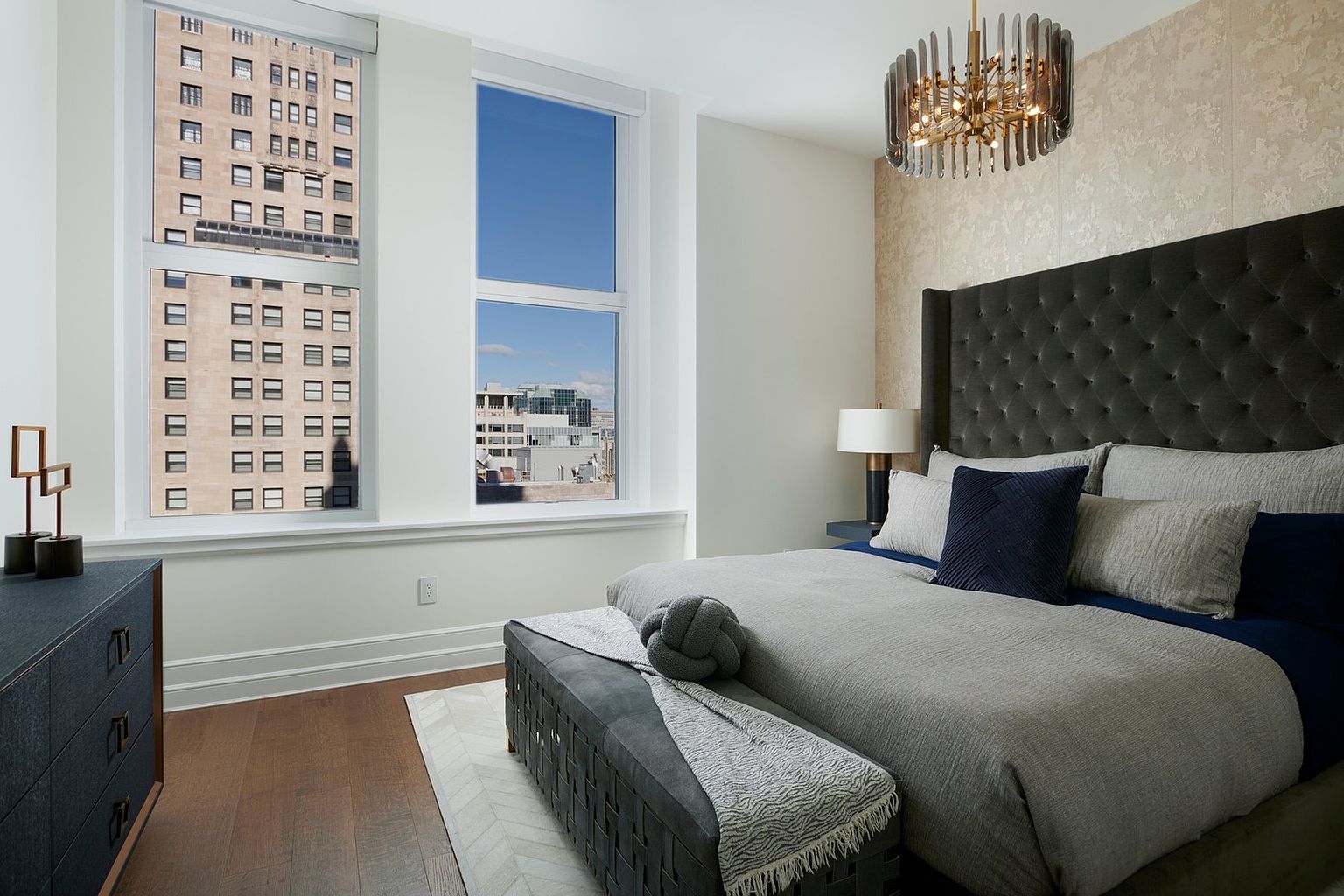 This sophisticated primary bedroom features a plush, tufted charcoal headboard set against a textured, neutral-toned wallpaper. The room is anchored by a grey upholstered bench at the foot of the bed and a modern chandelier, while large windows provide an expansive urban view. The overall aesthetic is one of refined, contemporary luxury with a focus on comfort and high-end finishes.