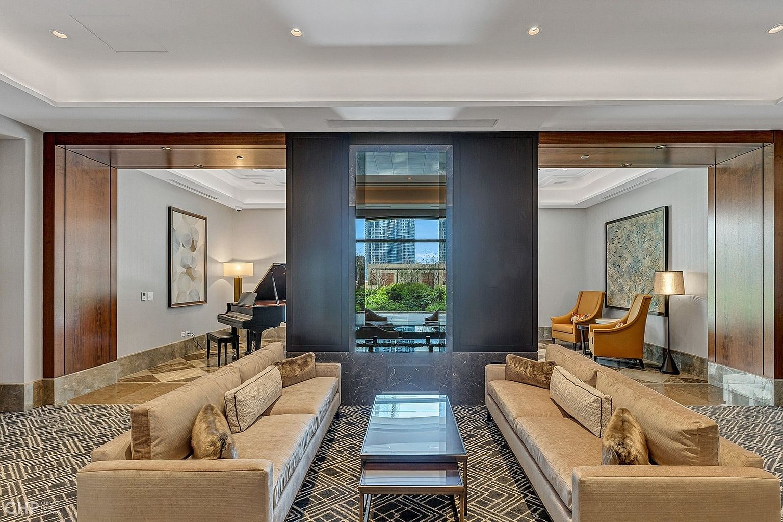 This sophisticated lounge area serves as a luxurious community amenity, featuring two plush, neutral-toned sofas facing a central glass coffee table. The space is anchored by a striking dark marble-clad wall with a central window looking out onto a landscaped courtyard, flanked by symmetrical seating areas that include a grand piano and elegant armchairs. The overall aesthetic is refined and upscale, characterized by high-end finishes, geometric patterned carpeting, and warm, ambient lighting.
