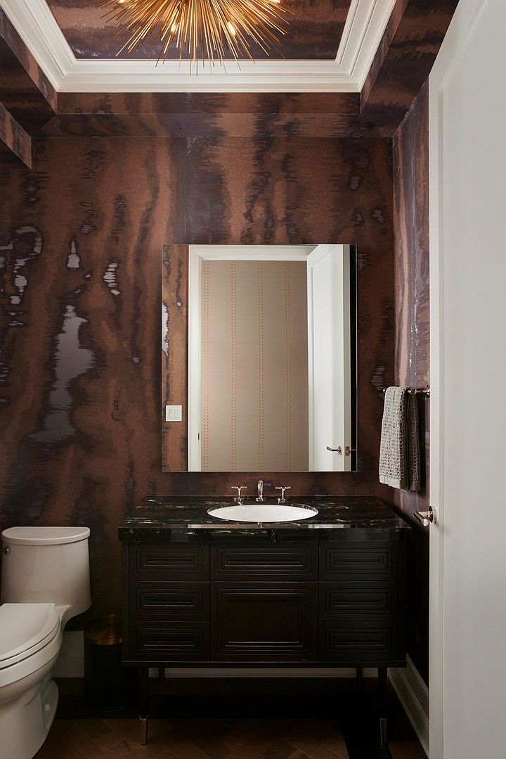 This sophisticated powder room features dramatic, dark, textured wallpaper that creates a moody and luxurious atmosphere. The space is anchored by a sleek black vanity with a dark stone countertop, a modern rectangular mirror, and a striking starburst chandelier overhead. The composition is centered and symmetrical, highlighting the high-end finishes and elegant design choices.