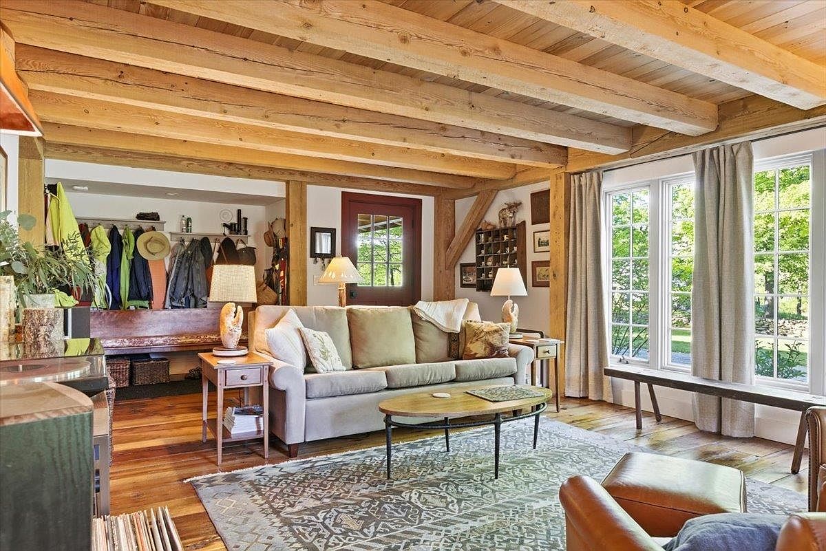 This cozy living room features exposed timber ceiling beams and warm wood flooring, creating a rustic and inviting atmosphere. A neutral-toned sofa sits centrally on a patterned area rug, complemented by a wooden coffee table and matching side tables with lamps. Large windows with light curtains allow natural light to fill the space, while an adjacent entryway area displays a coat rack and bench, highlighting the home's functional layout.